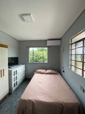 Una habitación con cama, ventana con vista a follaje, armario blanco y equipo de aire acondicionado.