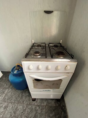 Una estufa a gas blanca con cuatro quemadores y un horno, ubicada en una esquina con un cilindro de gas azul a su lado.
