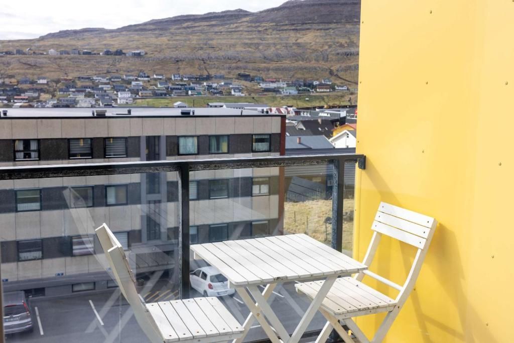 A balcony with a white folding table and chair, overlooking a town with buildings and a hill in the background.