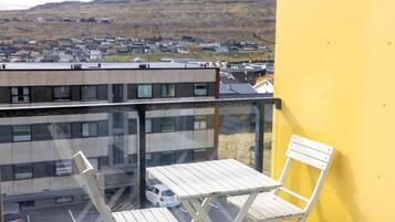 A balcony with a white folding table and chair, overlooking a town with buildings and a hill in the background.