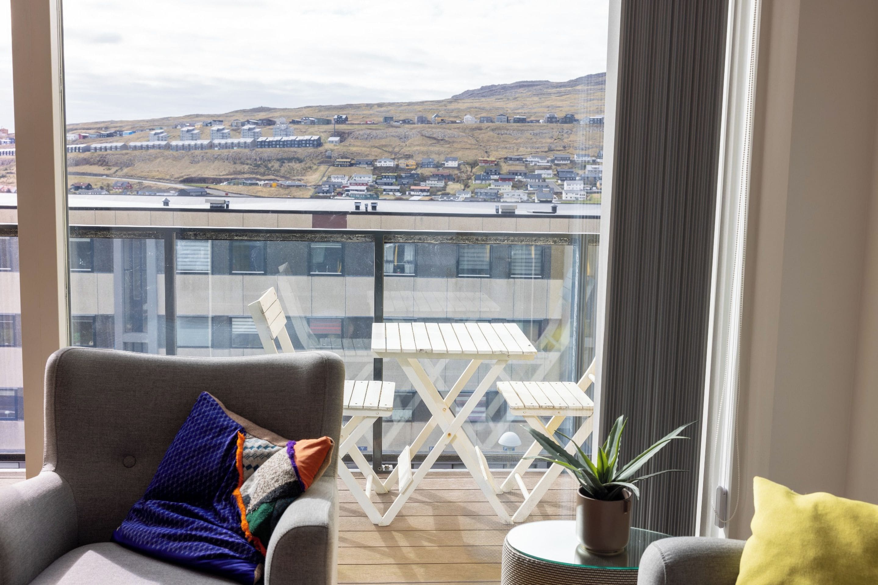A balcony with a view of a town, a grey sofa with a colorful cushion, and a small round table with a plant.