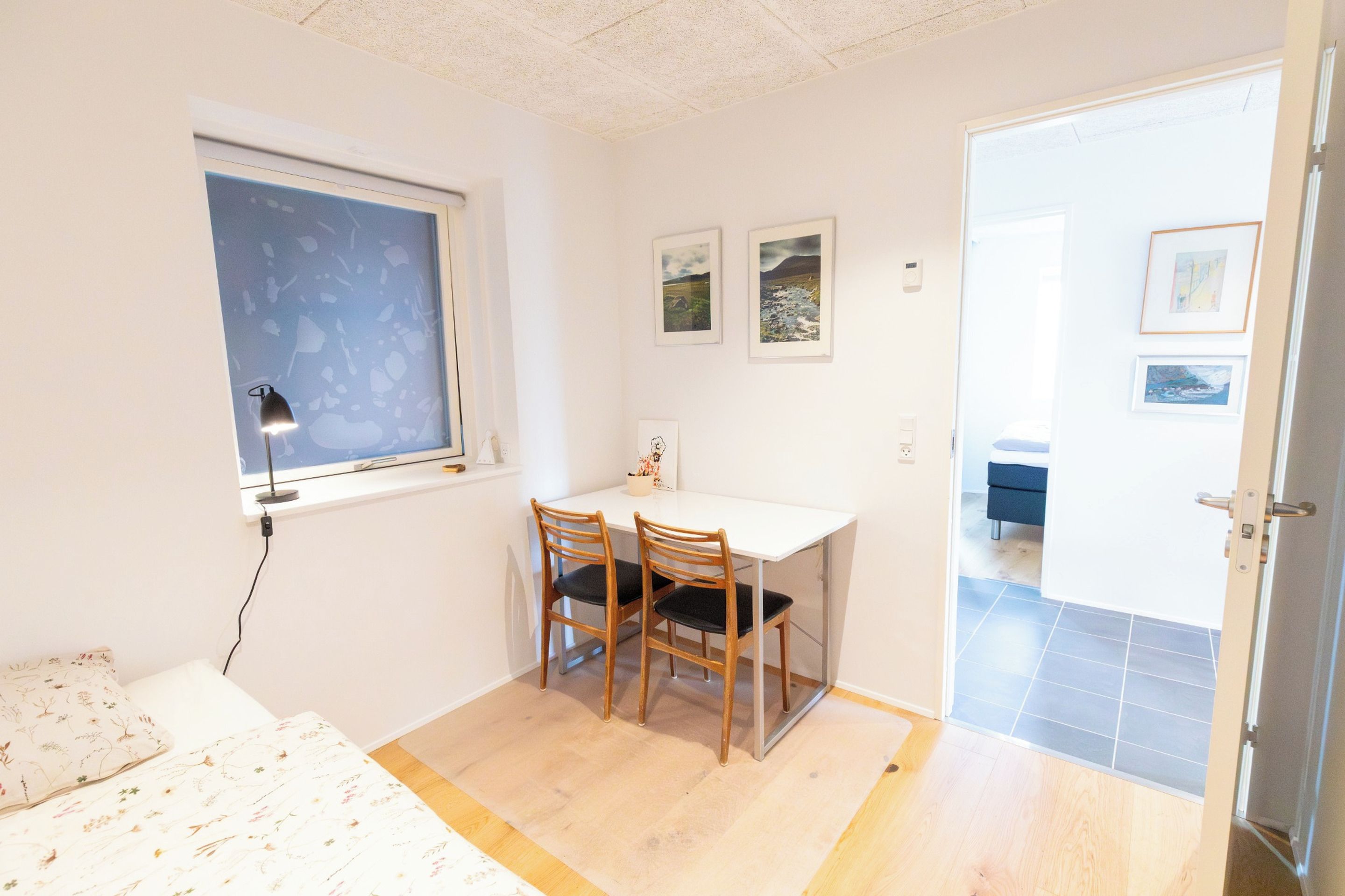 Una habitación pequeña y ordenada con un escritorio, dos sillas, una cama, un ventanal con vistas y una puerta que da a otra habitación.