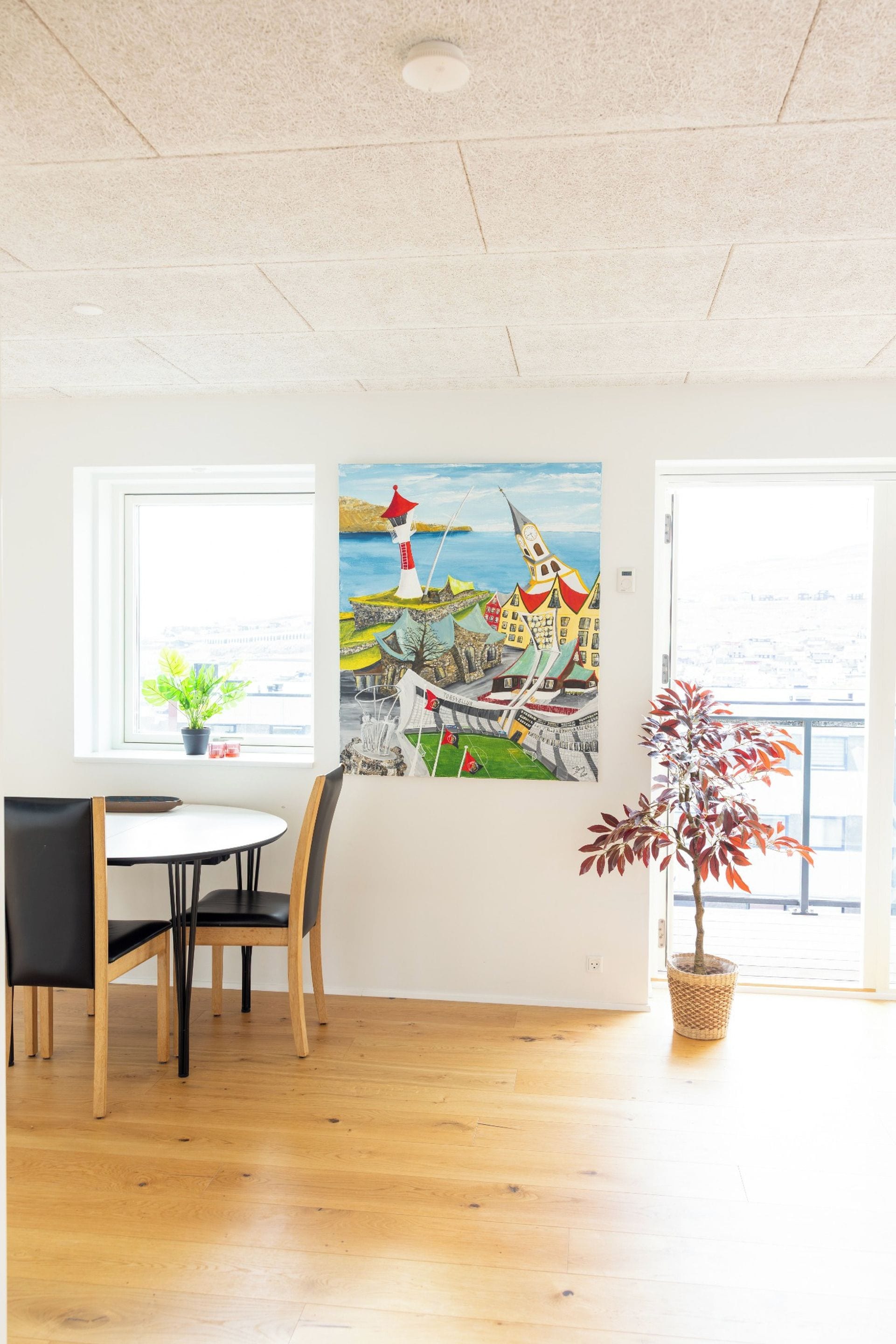 A room with a dining table, chairs, a potted plant, and a painting of a coastal scene.
