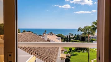 Une vue depuis une fenêtre donnant sur une zone côtière, avec un ciel dégagé, des palmiers et une vue lointaine sur la mer.