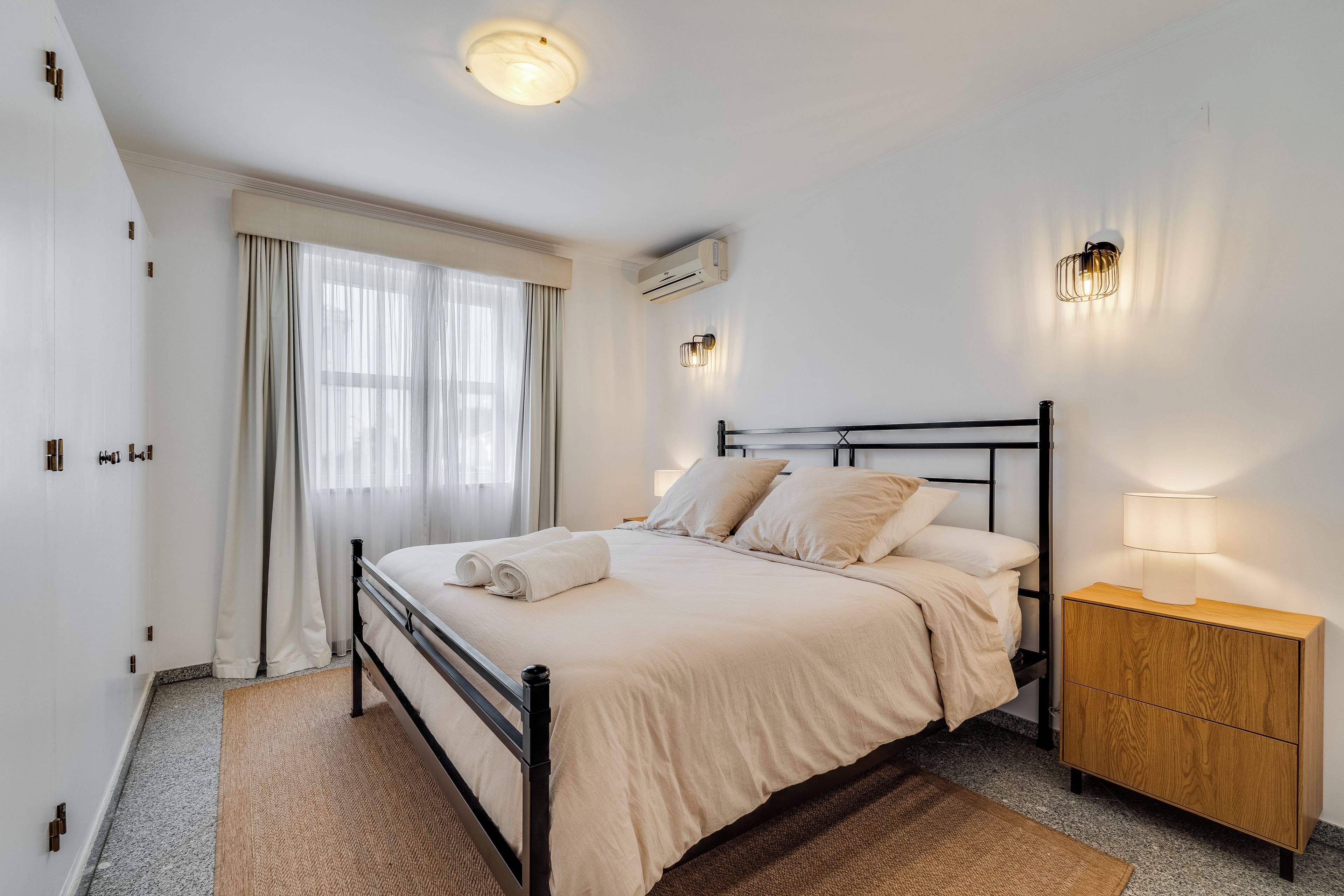 Habitació d'hotel amb llit, taulell de nit, làmpada, aparell d'aire condicionat i cortines.