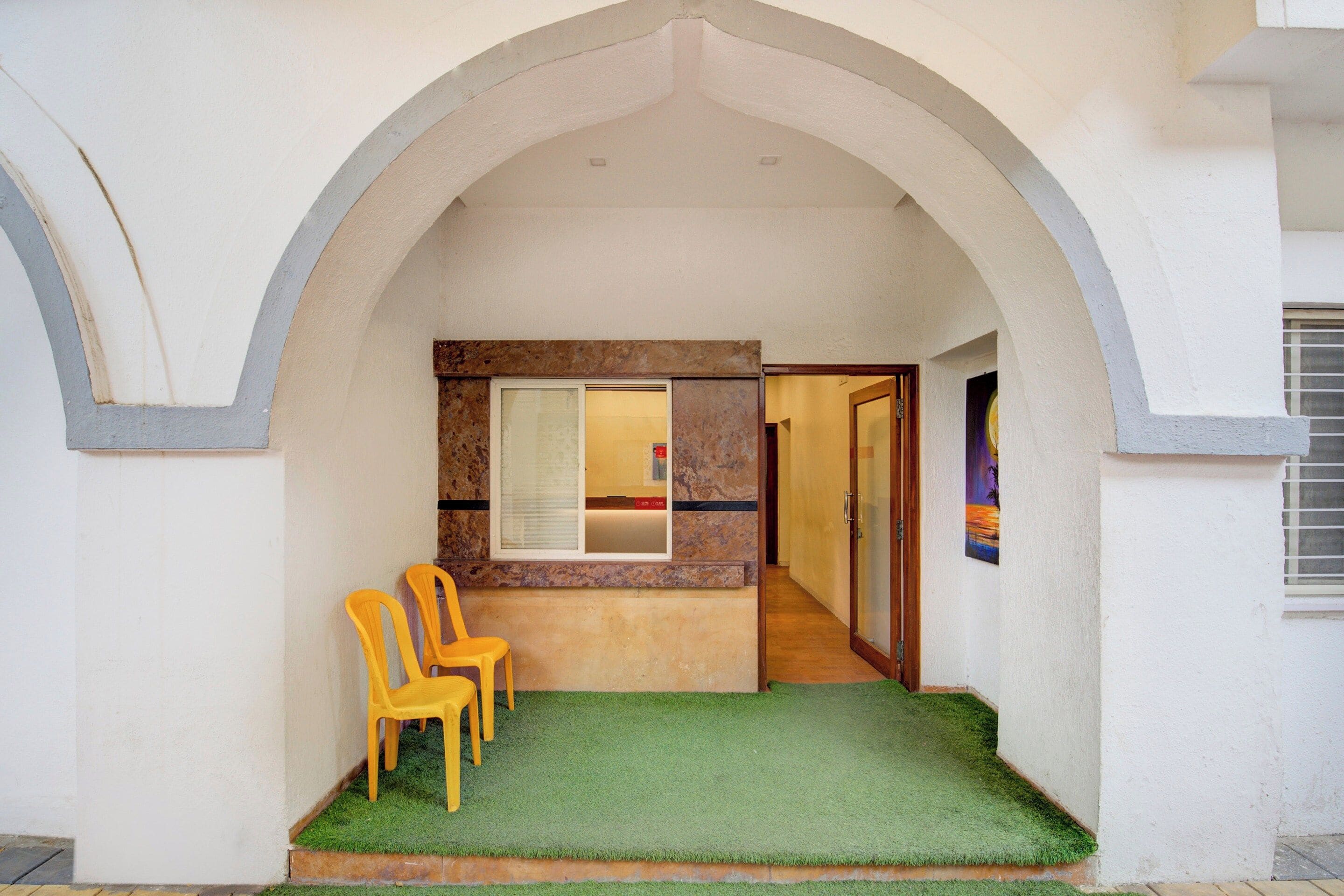 A covered outdoor area with a green artificial turf, two yellow chairs, and a stone wall with a window.