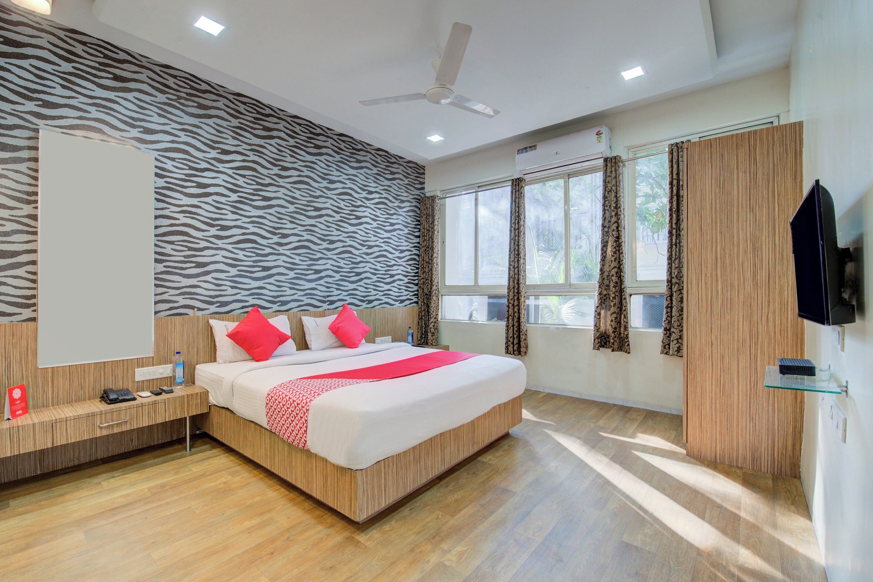 A modern hotel room with a zebra-patterned wall, a large bed with red and white bedding, a wooden floor, a ceiling fan, and a TV mounted on the wall.