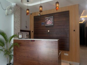 A reception desk with a wooden finish, a wall-mounted fan, and decorative lighting fixtures. - Classic Hotel C&D Stay (Indore)