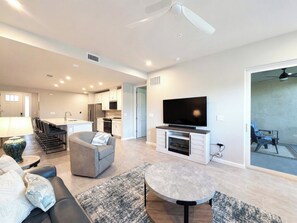 Una sala moderna con sofá, sillón y mesa de centro, televisor de pantalla plana y comedor con sillas, con una cocina al fondo.