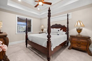 A four-poster bed with a wooden frame and white bedding, a ceiling fan, a window with blinds, and two bedside tables with lamps. - Coral Breeze Retreat (Cape Coral)