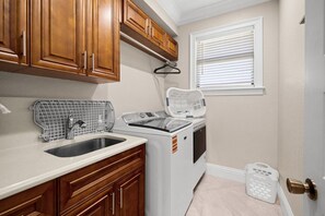 A kitchen with wooden cabinets, a stainless steel sink, a white washing machine, and a window with blinds. - Coral Breeze Retreat (Cape Coral)