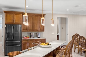 A modern kitchen with wooden cabinets, a stainless steel refrigerator, a white countertop, and hanging pendant lights. - Coral Breeze Retreat (Cape Coral)