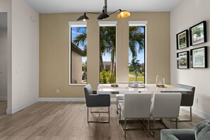 A modern dining area with a large table, chairs, pendant lighting, and large windows offering a view of palm trees and a house. - Terra Cove | Heated Pool, Dock & Gulf Access (Cape Coral)