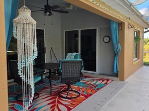 A covered patio area with a ceiling fan, a table with chairs, a decorative macramé hanging, and a colorful rug. - Lehigh Acres Cozy Retreat | Fort Myers Area (Lehigh Acres)