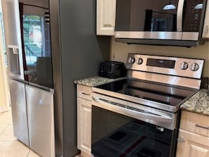 A compact kitchen with a stainless steel refrigerator, a built-in oven, and a microwave. - Lehigh Acres Cozy Retreat | Fort Myers Area (Lehigh Acres)