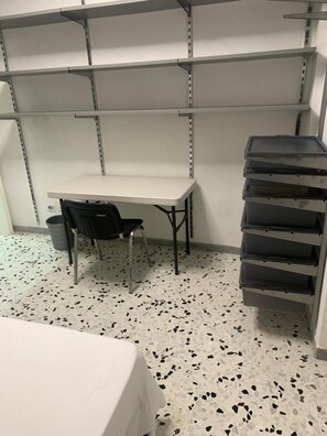 Een kamer met een witte tafel, een zwarte stoel en een stapel zwarte opbergbakken tegen een muur met metalen schappen.
