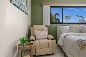 A bedroom with a bed, a recliner, a window with a view of palm trees, and a wall-mounted painting. - Punta Gorda Isles| Optional Guest  House (Punta Gorda)