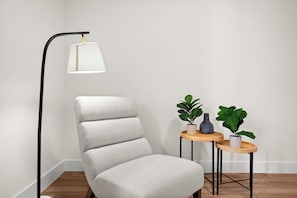 Canto de um quarto com uma poltrona estofada branca, um abajur e uma pequena mesa de madeira com duas plantas em vasos.