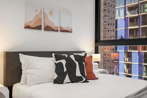 A hotel room with a bed, pillows, a nightstand, a lamp, and a window with a city view. - Level 34 City View 2 Bedroom (Melbourne)