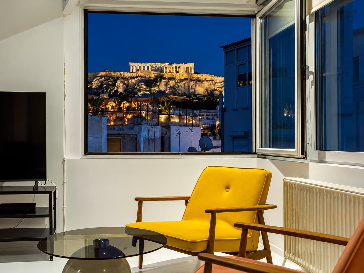 A room with a view of the Acropolis at sunset, a yellow armchair, a glass table, and a black TV stand.