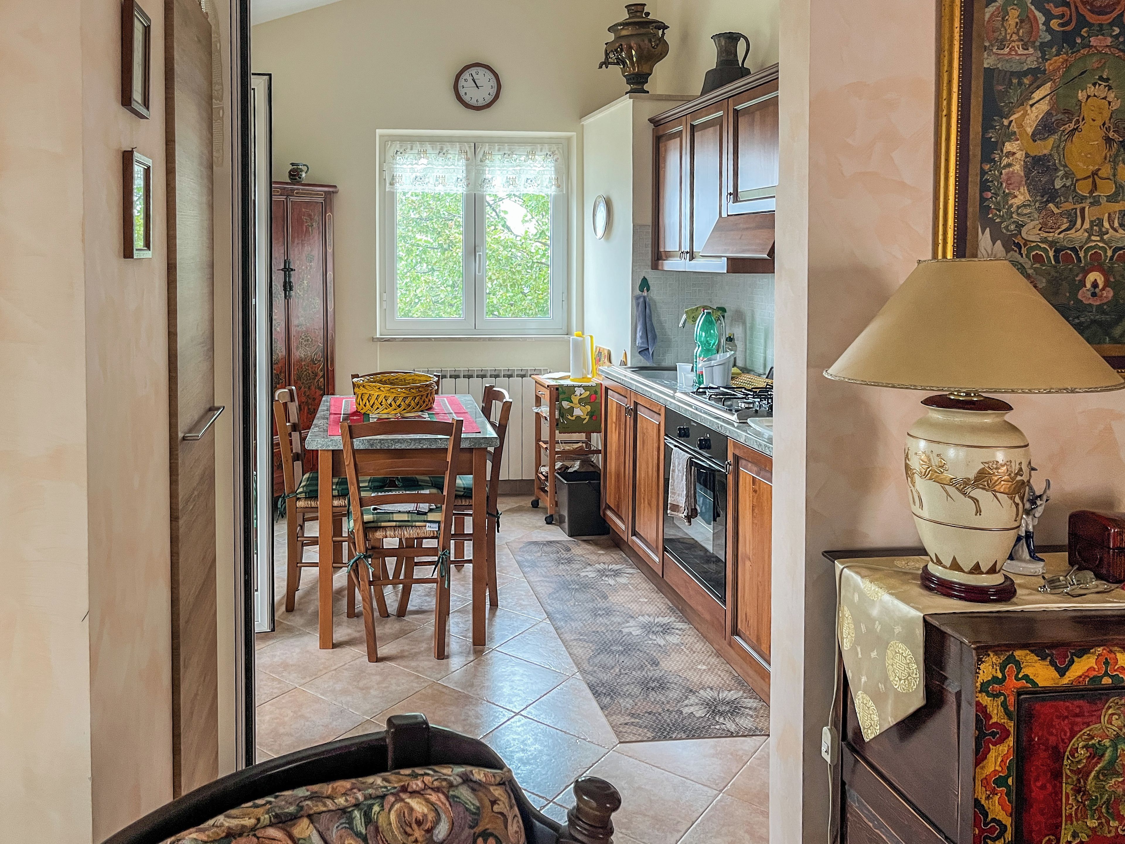 Une cuisine avec des meubles de rangement en bois, une table à manger avec des chaises, une fenêtre avec des rideaux et une lampe de table.