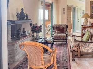 A living room with a fireplace, a patterned armchair, a rattan chair, and a coffee table. - Casa Censi F1 (Rocca Santo Stefano)