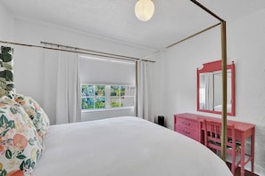 Ein Schlafzimmer mit einem Himmelbett, einem Fenster mit Vorhängen, einer rosa Kommode und einem Spiegel.