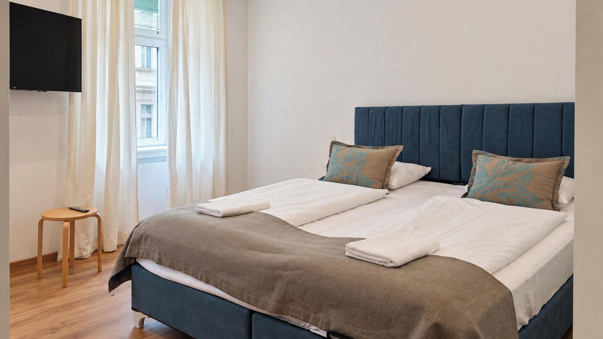 A hotel room with a bed, a TV mounted on the wall, a window with curtains, and a small stool.