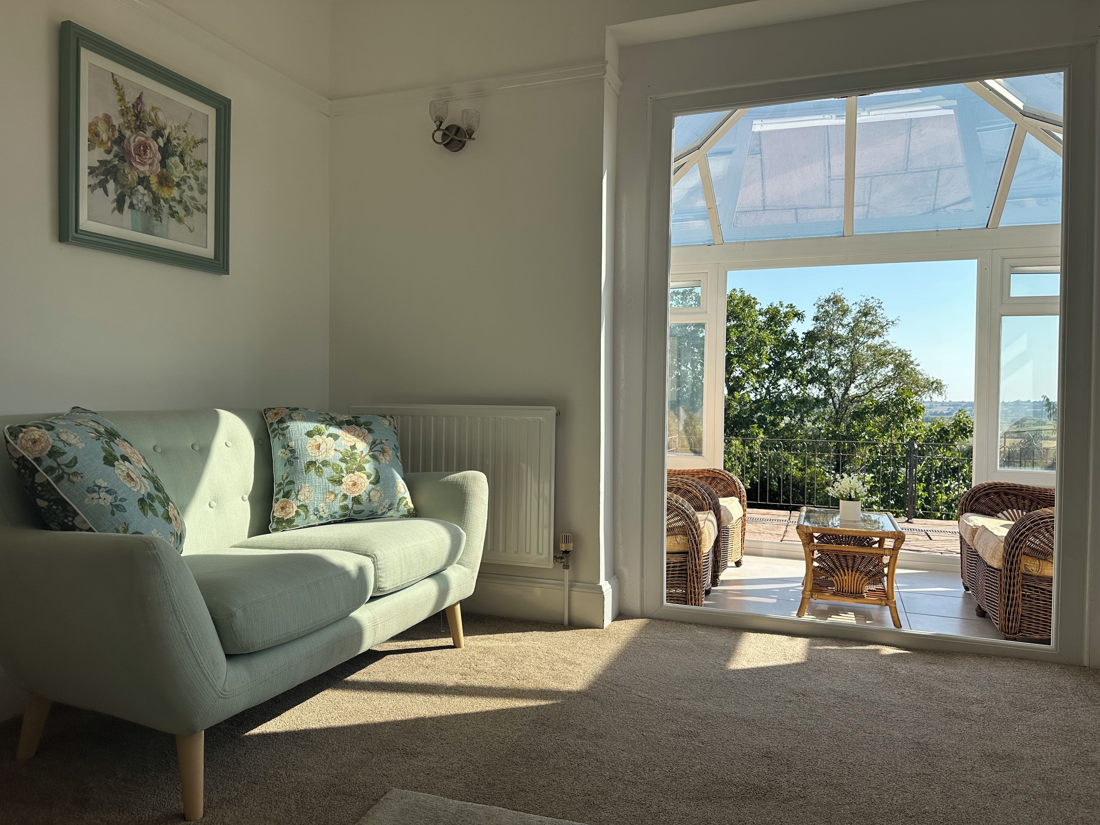 En stue med en lysegrøn sofa, blomsterpuder, et indrammet blomsterbillede og udsigt til en udendørs terrasse med fletmøbler.