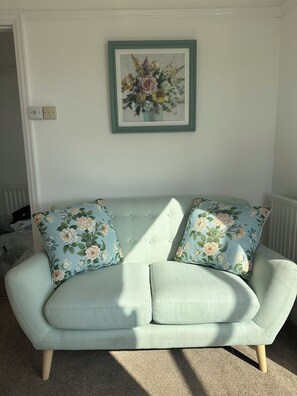 Un sofà verd clar amb coixins florals, un quadre floral emmarcat a la paret i un radiador a la dreta.