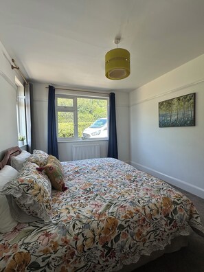 Een slaapkamer met een bed, een raam met gordijnen, een radiator, een plafondventilator en een schilderij aan de muur.