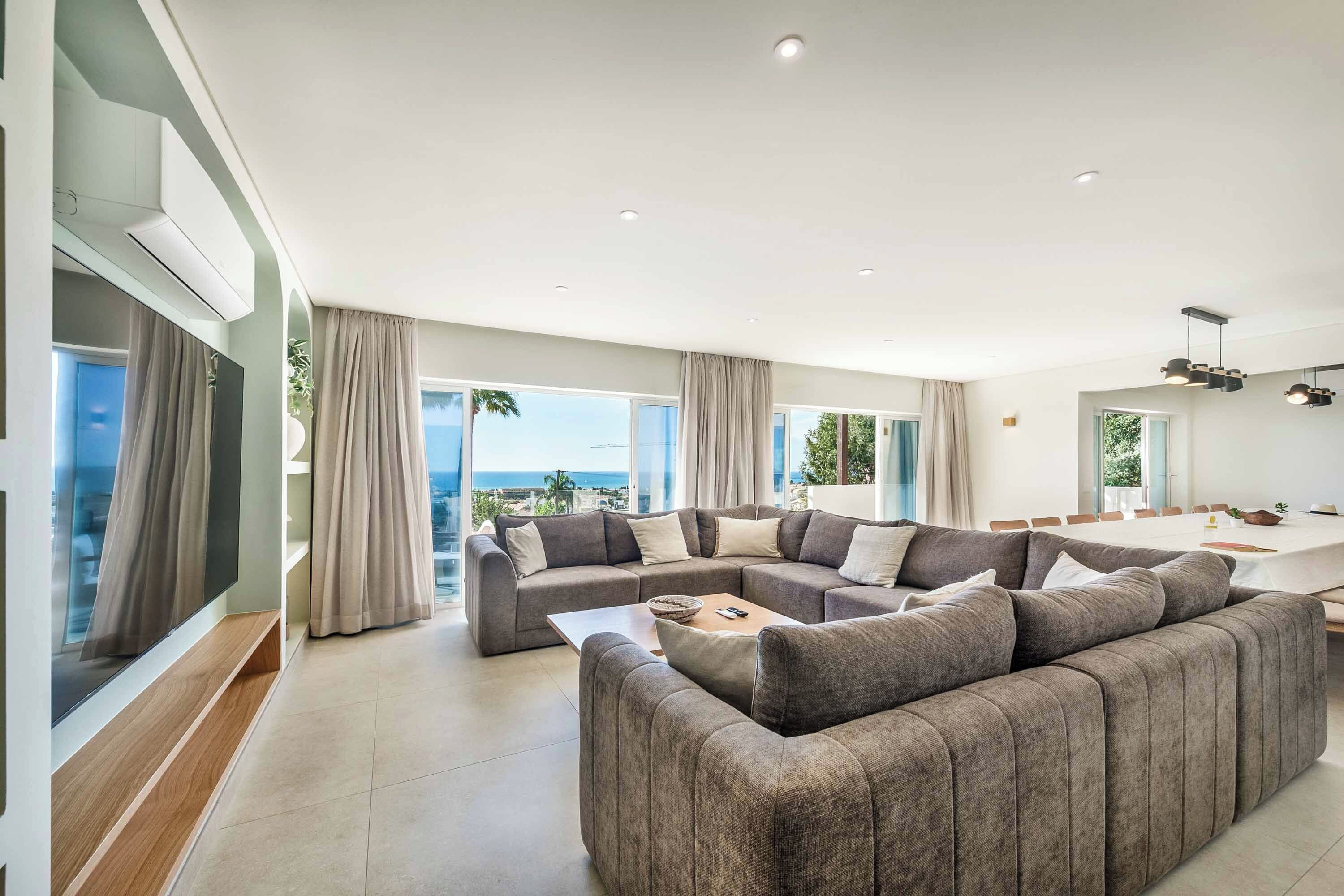 A modern living room with a sectional sofa, a dining area, and large windows offering a view of the sea.