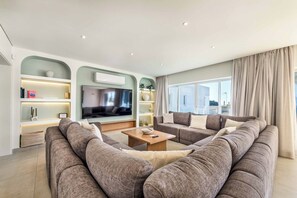 A modern living room with a sectional sofa, a flat-screen TV, built-in shelving, and a large window with curtains. - Villa Gardenia Vista (Albufeira)