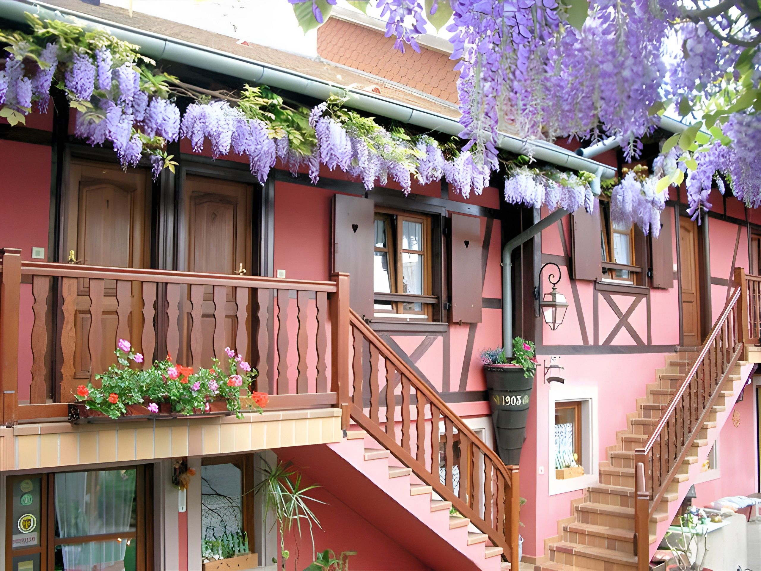 Un bâtiment doté d’un balcon, d’un escalier en bois et de fleurs violettes suspendues.
