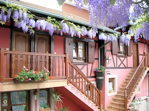 Un edificio con balcón, escalera de madera y flores moradas colgantes.