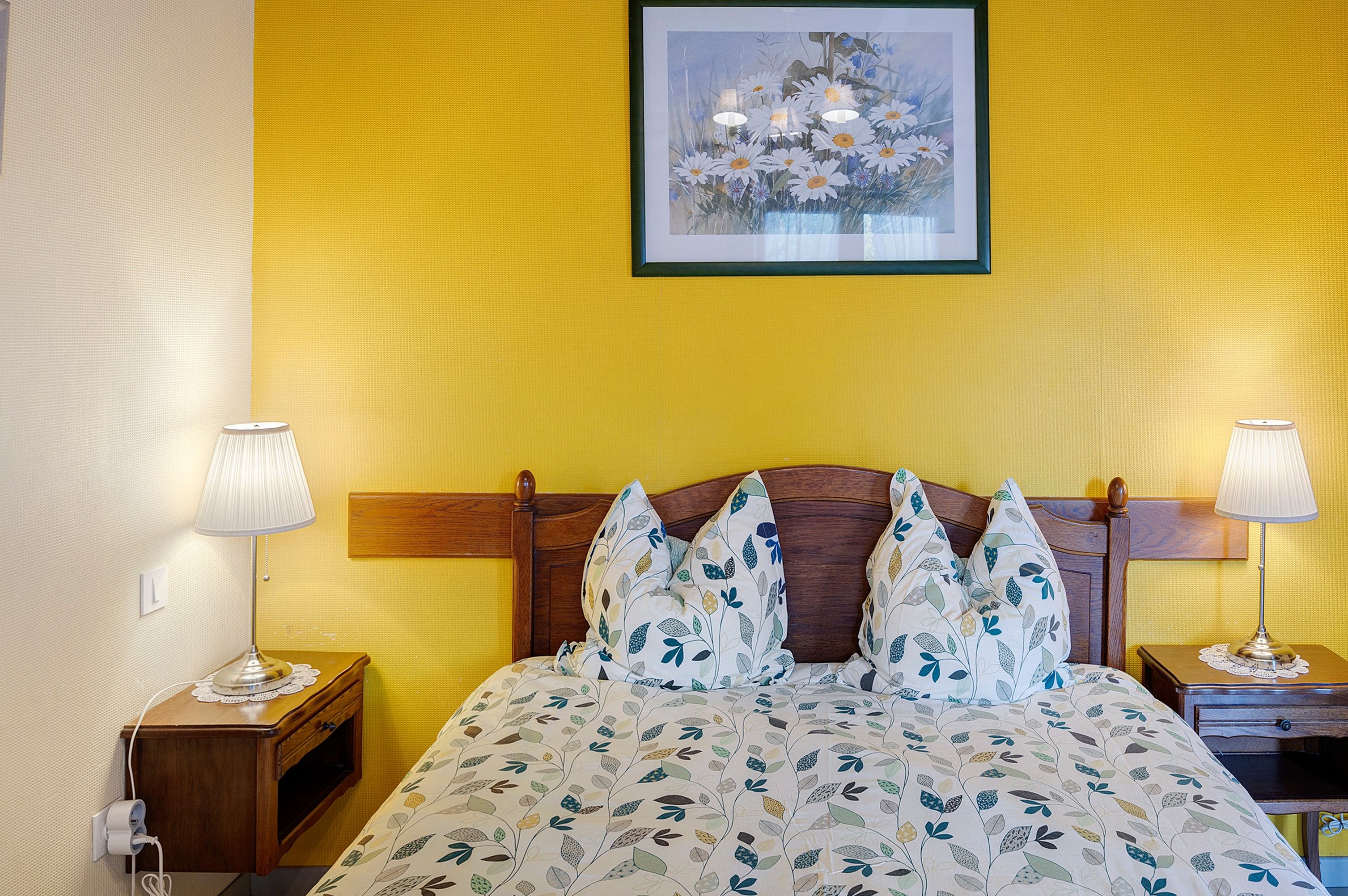 A hotel room with a bed, two bedside lamps, a wooden headboard, a wall-mounted picture, and a textured yellow wall.
