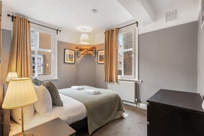Quarto com cama, mesa de cabeceira, abajur, radiador e janela com cortinas.