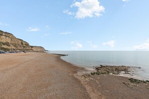 A pebbled beach with a calm sea, a cliff on the left, and a clear sky. - Hastings Buzz | By My Getaways (Hastings)