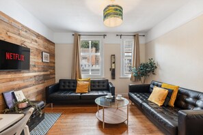A modern living room with two black leather sofas, a glass coffee table, and a wooden wall with a mounted television. - Hastings Buzz | By My Getaways (Hastings)