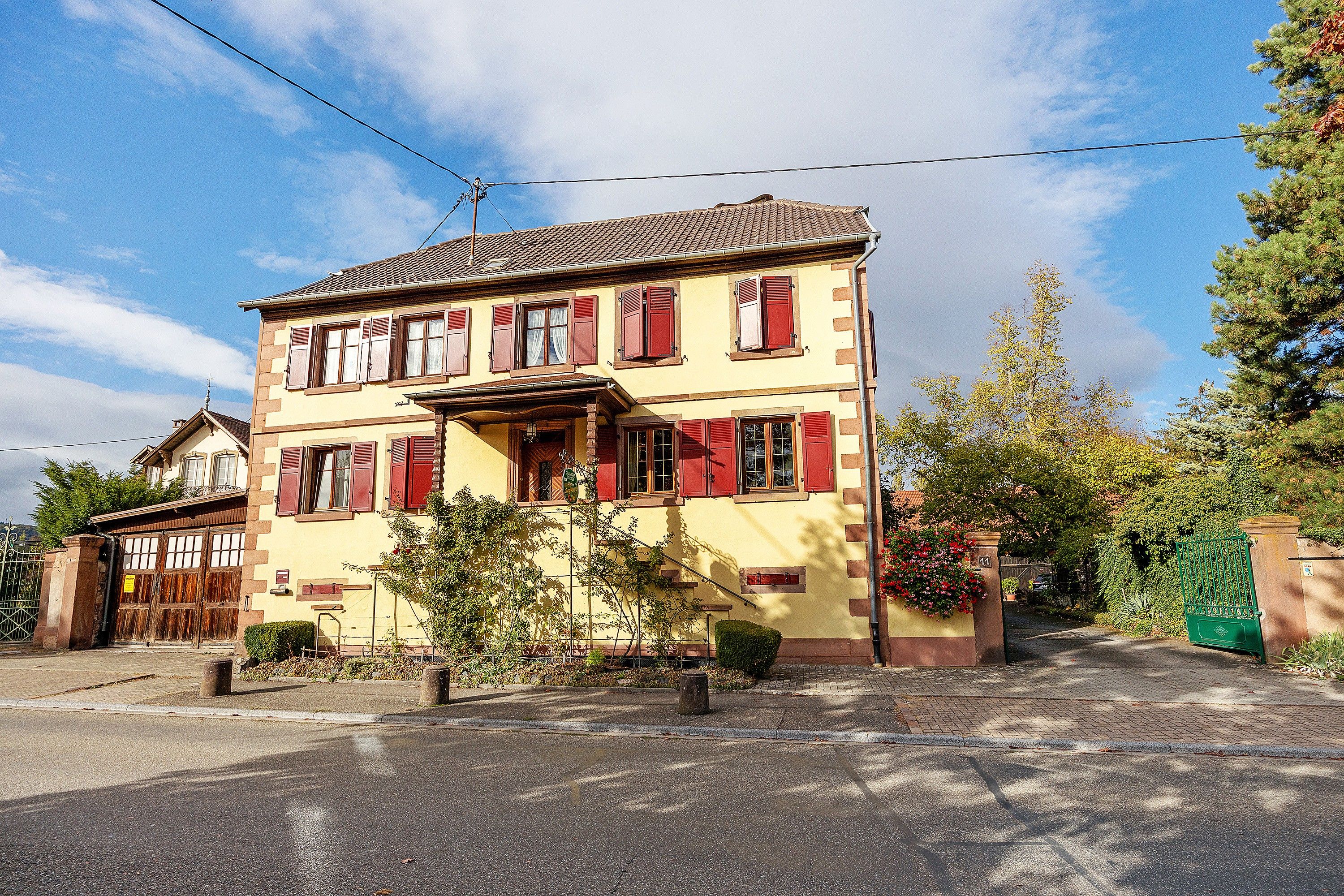 Un bâtiment à deux étages, avec une façade jaune et des volets rouges, une allée d’accès fermée par un portail et un ciel dégagé.