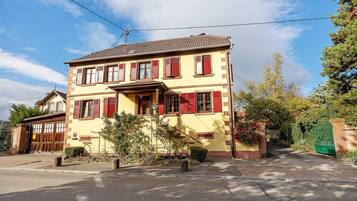 Un bâtiment à deux étages, avec une façade jaune et des volets rouges, une allée d’accès fermée par un portail et un ciel dégagé.
