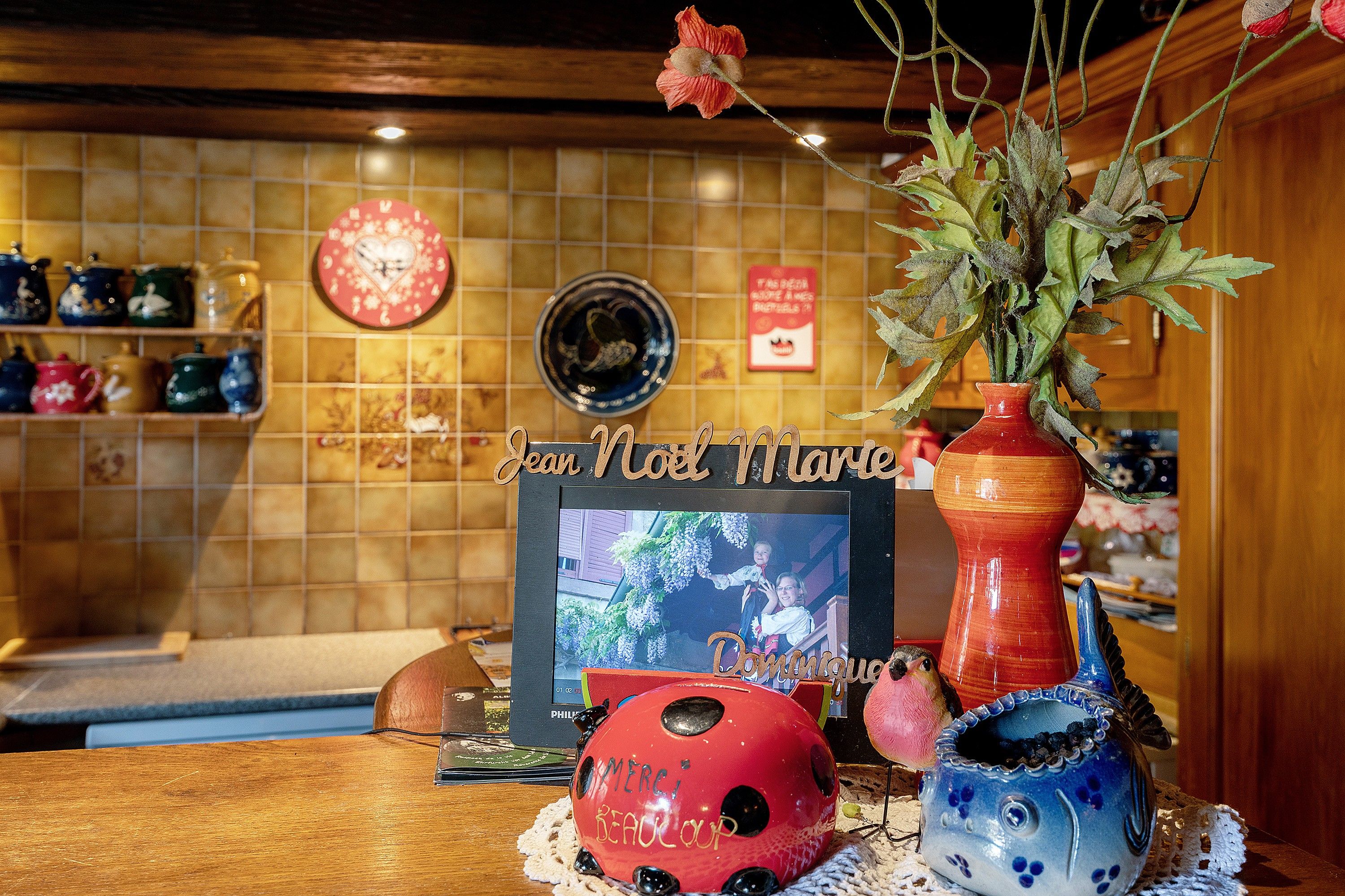 A room with a wooden wall, a clock, a framed photo, and a vase with dried flowers.