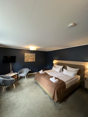 Ein Hotelzimmer mit einem Bett, zwei Sesseln, einem Schreibtisch, einem Flachbildfernseher und einem Bild an der Wand.