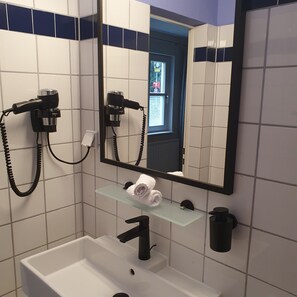 Ein Badezimmer mit einem weißen Waschbecken, einem schwarzen Wasserhahn und einer Glasablage mit einem Handtuch. Zwei Haartrockner sind an der Wand montiert, und über dem Waschbecken hängt ein Spiegel.