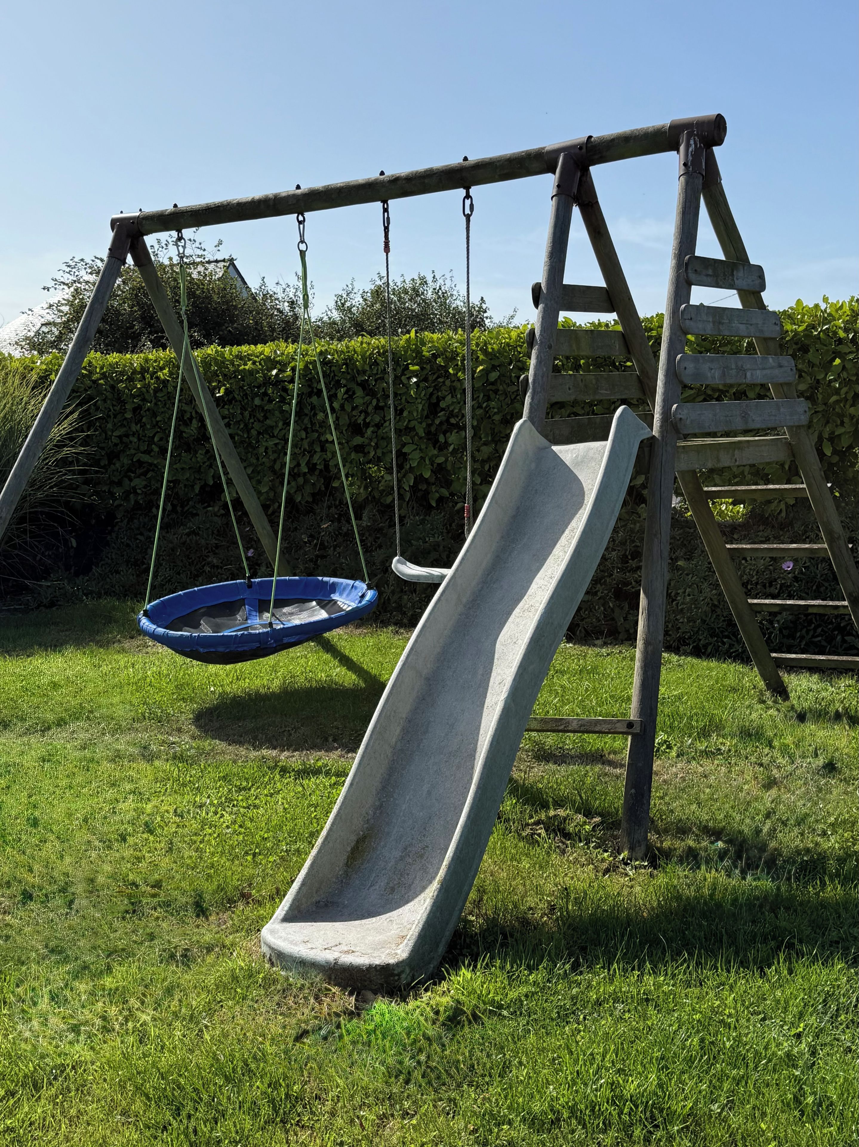 Un ensemble de jeux d’extérieur en bois, comprenant un toboggan et une balançoire suspendue, installé dans un espace herbeux.