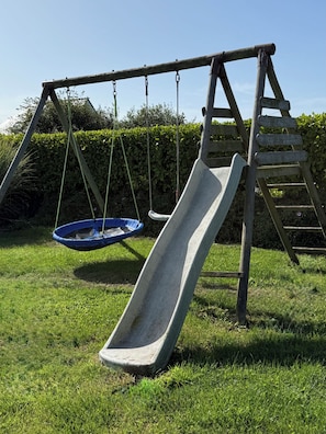 A wooden swing set with a slide and a hanging swing in a grassy area. - Casa Isann—Pool—Ocean View & GR34 (Landéda)