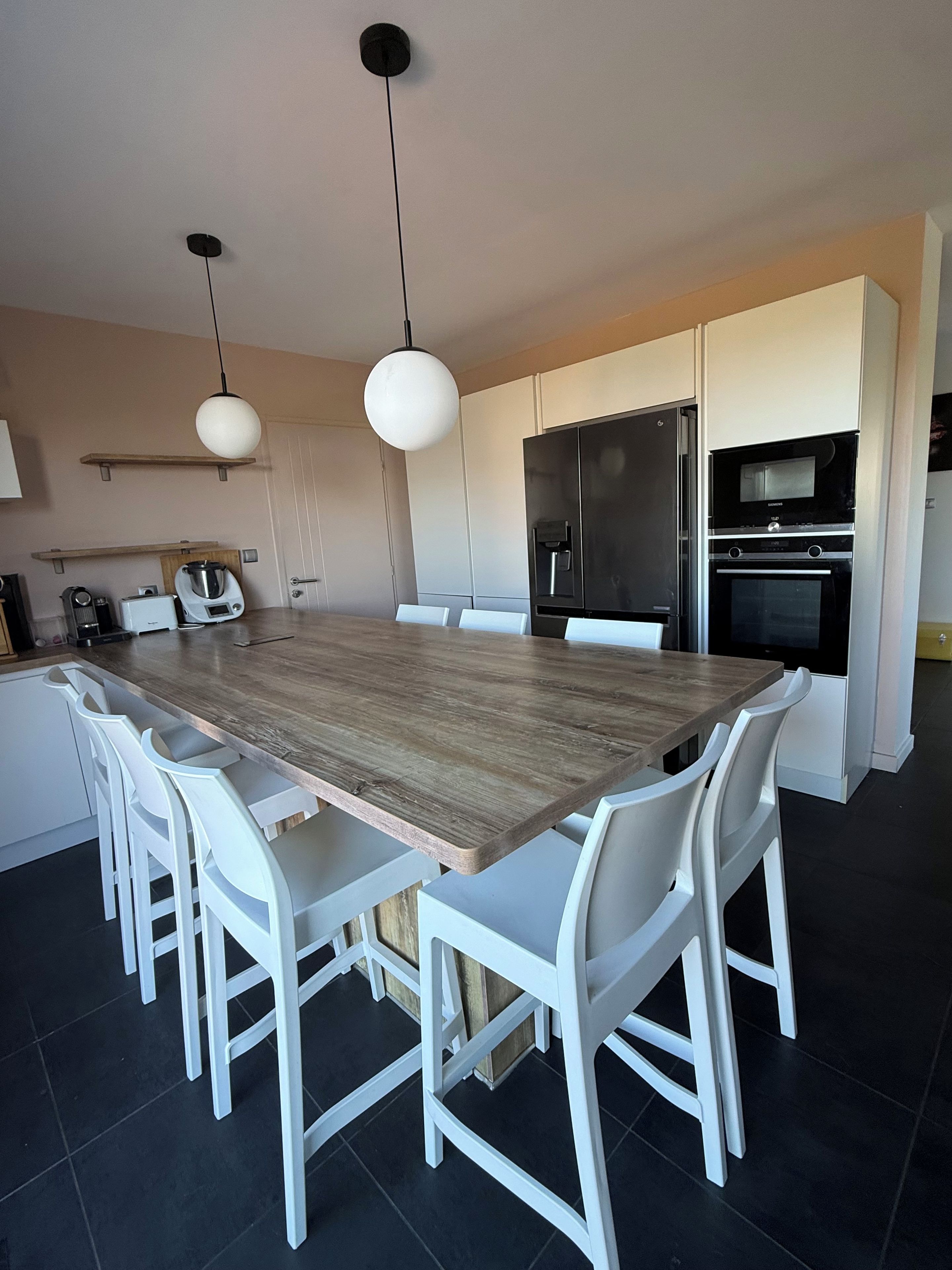 Een moderne keuken met een grote houten tafel, witte stoelen en ingebouwde apparatuur, waaronder een koelkast en oven.
