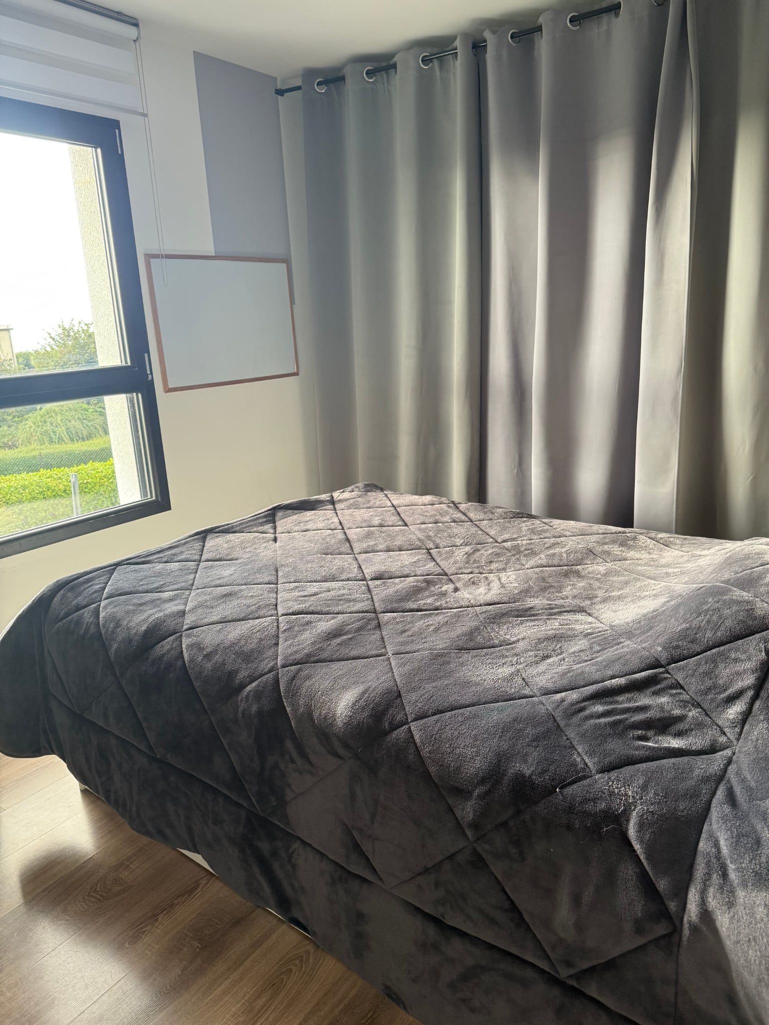 Ein Schlafzimmer mit einem Bett, einem Fenster mit Vorhängen und Blick auf Grünfläche.