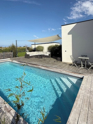 A swimming pool with a wooden deck, a seating area with cushions, and a white wall. - Vrbo Property (Landéda)