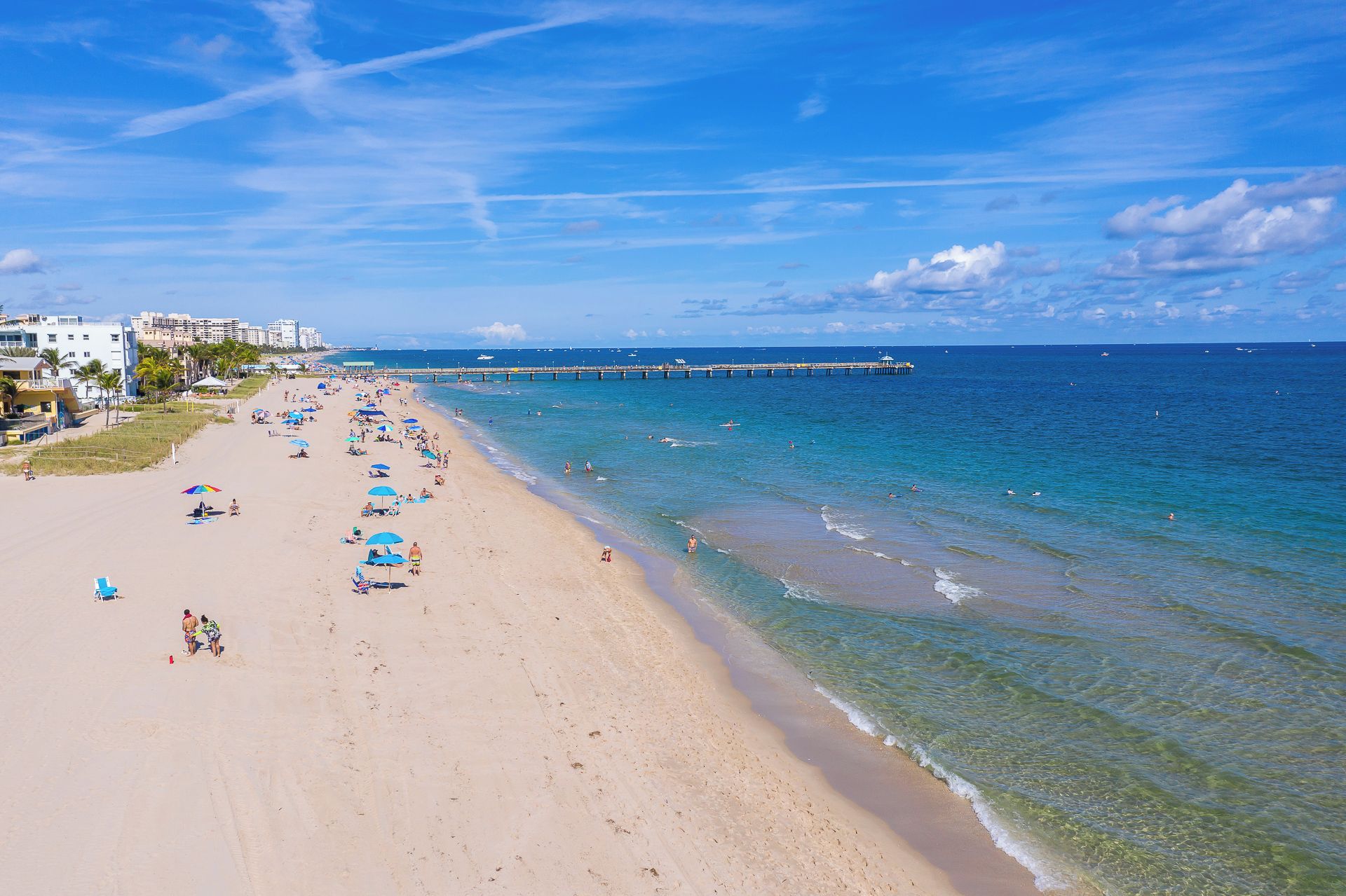 A beach with clear blue water, a pier, and buildings in the background.